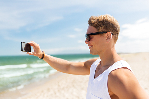 summer holidays and people concept - happy smiling young man with smartphone on beach photographing sea