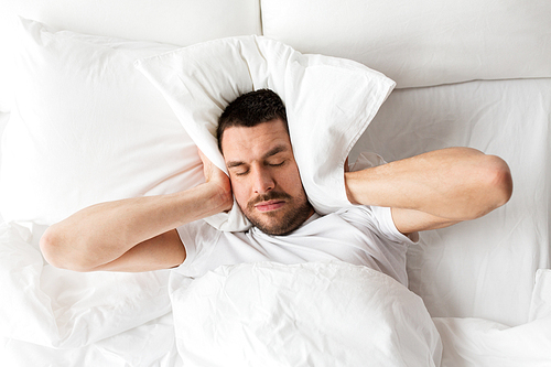 people, bedtime and rest concept - man lying in bed with pillow suffering from noise at home