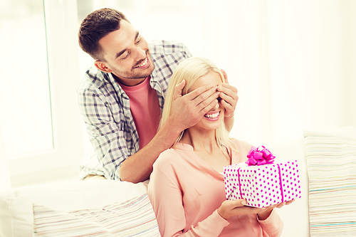 relationships, love, people, birthday and holidays concept - happy man covering womans eyes and giving gift box at home