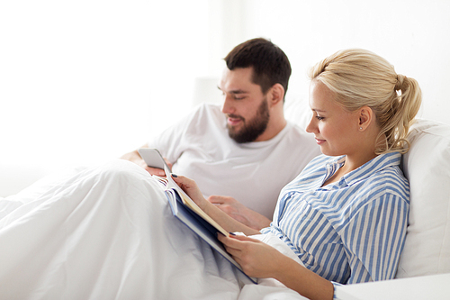 people, technology, internet, relations and communication concept - happy couple with book and smartphone in bed at home