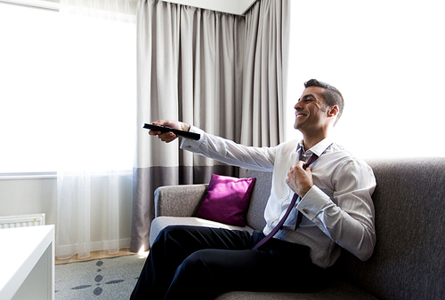 people and rest concept - happy businessman with tv remote taking off his tie at hotel room