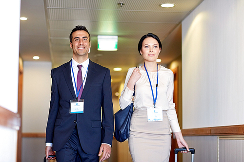 business trip and people concept - man and woman with travel bags and conference badges at hotel corridor