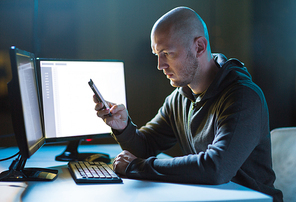 cybercrime, hacking and technology concept - male hacker with smartphone and coding on computers screen in dark room