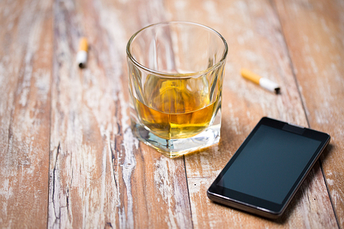 alcohol addiction and alcoholism concept - close up of glass of whiskey, smartphone and cigarette butts on table