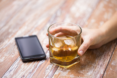 alcoholism, alcohol addiction and people concept - close up of male hand with glass of whiskey and smartphone on table