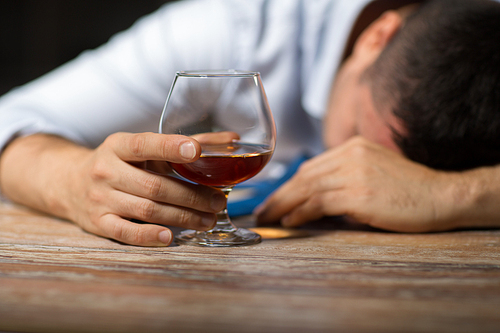 alcoholism, alcohol addiction and people concept - male alcoholic with glass of brandy lying or sleeping on table at night