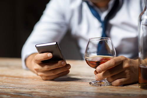 alcoholism, alcohol addiction and technology concept - close up of male hands with smartphone and glass of brandy