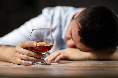 alcoholism, alcohol addiction and people concept - male alcoholic with glass of brandy lying or sleeping on table at night