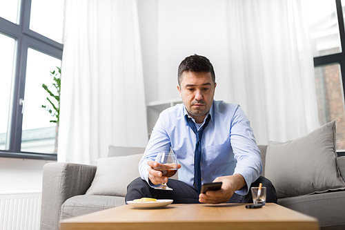 alcoholism, alcohol addiction and people concept - drunk man with smartphone and glass of brandy at home