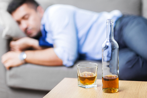 alcoholism, alcohol addiction and people concept - bottle with glass of whiskey on table and sleeping drunk man