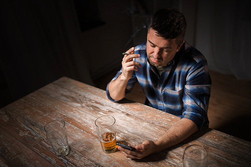 alcoholism, alcohol addiction and people concept - male alcoholic with smartphone drinking beer and smoking cigarette at night