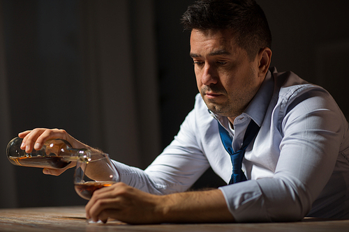 alcoholism, alcohol addiction and people concept - male alcoholic pouring brandy from bottle to glass at table at night