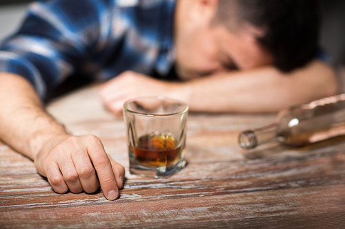 alcoholism, alcohol addiction and people concept - male alcoholic with glass of whiskey and bottle lying or sleeping on table at night