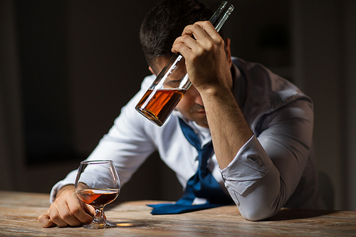 alcoholism, alcohol addiction and people concept - male alcoholic with bottle drinking brandy at table at night