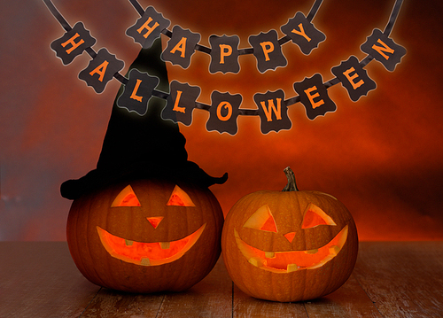 holidays, decoration and celebration concept - jack-o-lanterns or pumpkins in witch hat and happy halloween festive garland or banner over dark background