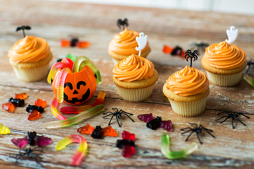 food, baking and holidays concept - cupcakes or frosted muffins with halloween party decorations and candies on wooden table