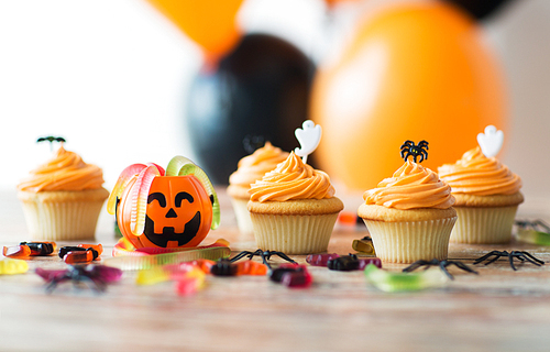 food, baking and holidays concept - cupcakes or frosted muffins with halloween party decorations and candies on wooden table