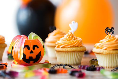 halloween and holidays concept - cupcakes or frosted muffins with party decorations and candies on wooden table