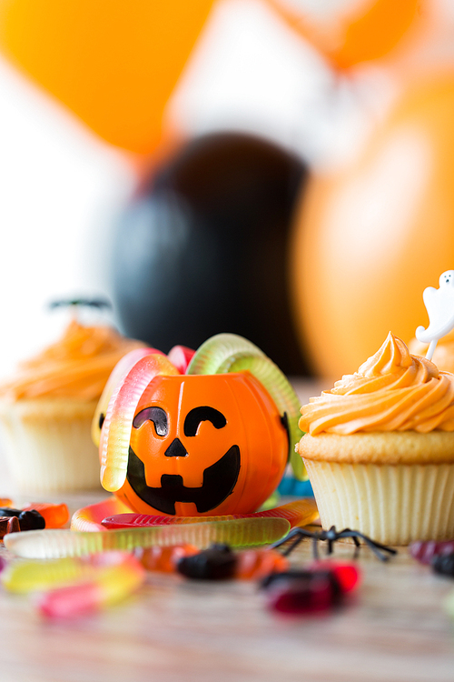 halloween and holidays concept - cupcakes or frosted muffins with party decorations and candies on wooden table