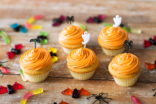 food, baking and holidays concept - cupcakes or frosted muffins with halloween party decorations and candies on wooden table