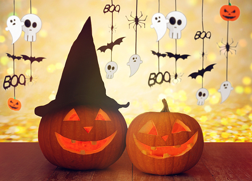 holidays, decoration and celebration concept - jack-o-lantern or pumpkins in witch hat and halloween festive garland over lights background