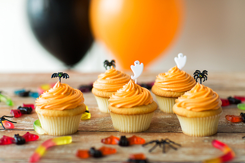 food, baking and holidays concept - cupcakes or muffins with halloween party decorations and jelly candies on wooden table and balloons