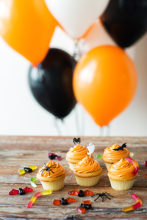food, baking and holidays concept - cupcakes or muffins with halloween party decorations and jelly candies on wooden table and balloons