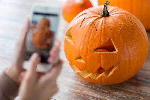 halloween, decoration and holidays concept - close up of jack-o-lantern or carved pumpkin and hands with smartphone