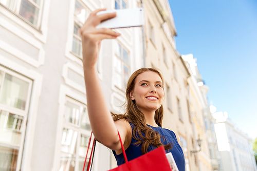sale, consumerism, technology and people concept - happy young woman with shopping bags and smartphone taking selfie on city street
