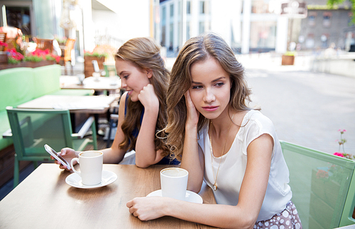 technology, internet addiction and people concept - young woman using smartphone and sad friend drinking coffee at cafe outdoors