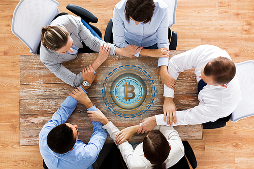cryptocurrency, cooperation and finance concept concept - business team sitting at office table with bitcoin hologram and holding hands from top