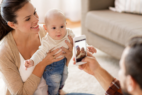 family, parenthood and people concept - happy father with smartphone taking picture of mother with baby at home