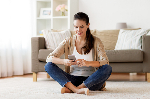 people, technology, communication and leisure concept - happy young woman sitting on floor and texting message on smartphone at home