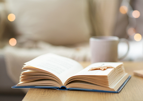 hygge, literature and reading concept - open book with autumn leaf on wooden table at home