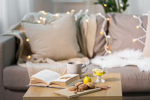 hygge and cozy home concept - oatmeal cookies, book, tea and lemon on wooden table in living room