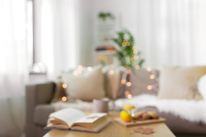 hygge and cozy home concept - blurred background of living room with book, cup of tea and food on table and sofa
