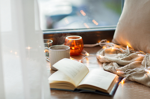 hygge and cozy home concept - book, cup of coffee or hot chocolate and candles with garland on window sill