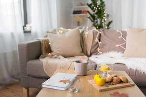 hygge and cozy home concept - cup of tea with lemon, book, autumn leaves and oatmeal cookies on wooden table in living room