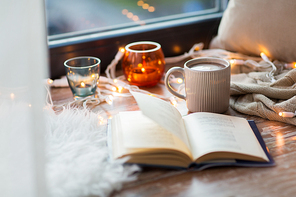 hygge and cozy home concept - book, cup of coffee or hot cchocolate and candles with garland on window sill