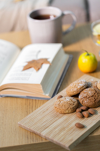 hygge, bake and food concept - oatmeal cookies, almonds, book and tea on wooden table at home