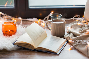 hygge and cozy home concept - book, cup of coffee or hot chocolate and candles with garland on window sill