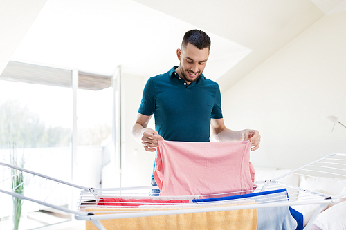 laundry and household concept - happy man taking clothes from drying rack and putting them to basket at home