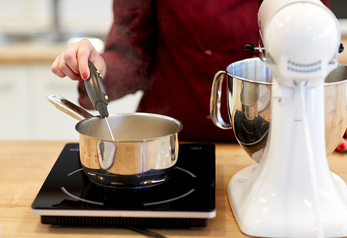 cooking, food and kitchen appliances concept - chef with stand mixer and thermometer measuring temperature in sugar syrup boiling in pot on stove at kitchen