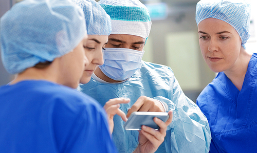 surgery, medicine and people concept - group of surgeons with smartphone in operating room at hospital