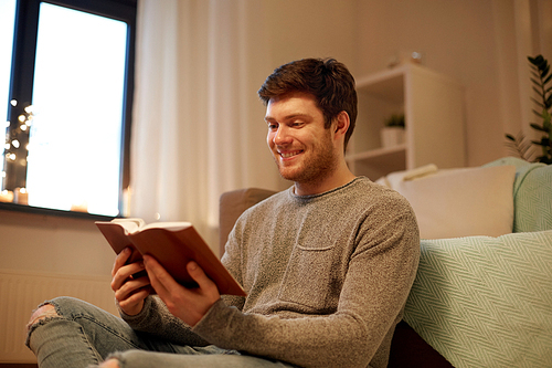 leisure, hygge and people concept - happy young man reading book at home