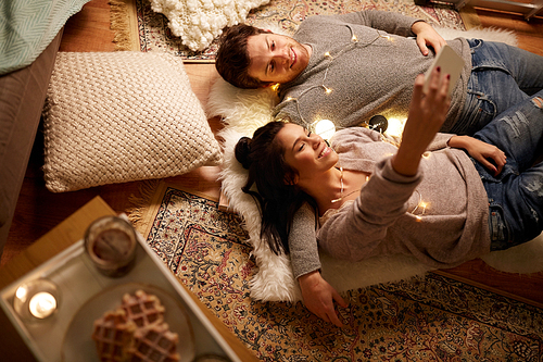 leisure, hygge and people concept - happy couple with garland lying on floor at home and making selfie by smartphone
