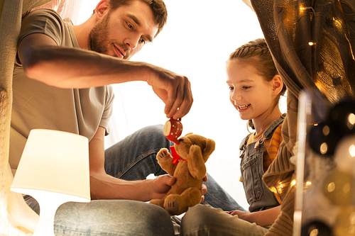 family, hygge and people concept - happy father and little daughter playing with teddy bear and toy tea pot in kids tent at night at home