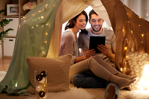 leisure, hygge, technology and people concept - happy couple with tablet pc computer in kids tent at home
