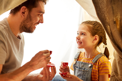 family, hygge and people concept - happy father and little daughter playing tea party in kids tent at night at home