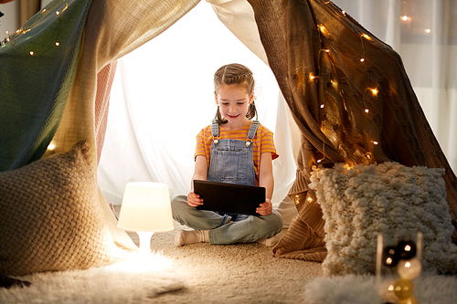 childhood, technology and hygge concept - happy little girl with tablet pc computer in kids tent at home